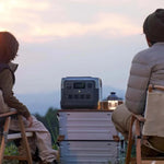 Two people sitting on chairs in front of a portable power station with a scenic background. Ecoflow