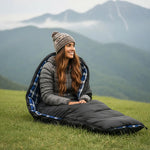 Woman sitting in a sleeping bag on a grassy field with mountains in the background