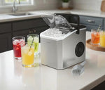 A stainless steel countertop ice bucket with a clear lid, filled with ice, shown on a kitchen counter with glasses of iced drinks and a scoop nearby.