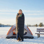 Person wrapped in a sleeping bag holding a mug in front of a tent in a snowy landscape