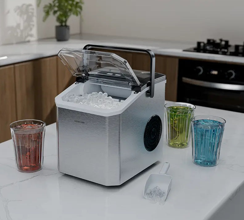 A stainless steel countertop ice machine with its lid open, filled with clear ice, shown on a white kitchen counter with three colored drinking glasses and a small scoop.