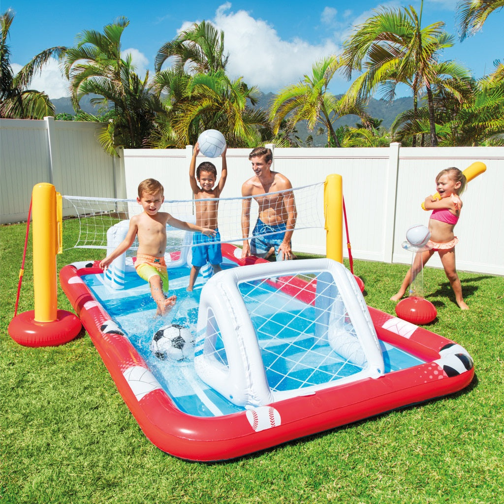 Children playing with an inflatable pool and sports set on a grassy area with palm trees in the background.