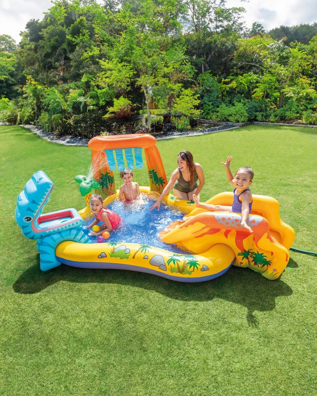 Children playing in an inflatable dinosaur-themed pool set on a grassy area with trees in the background.