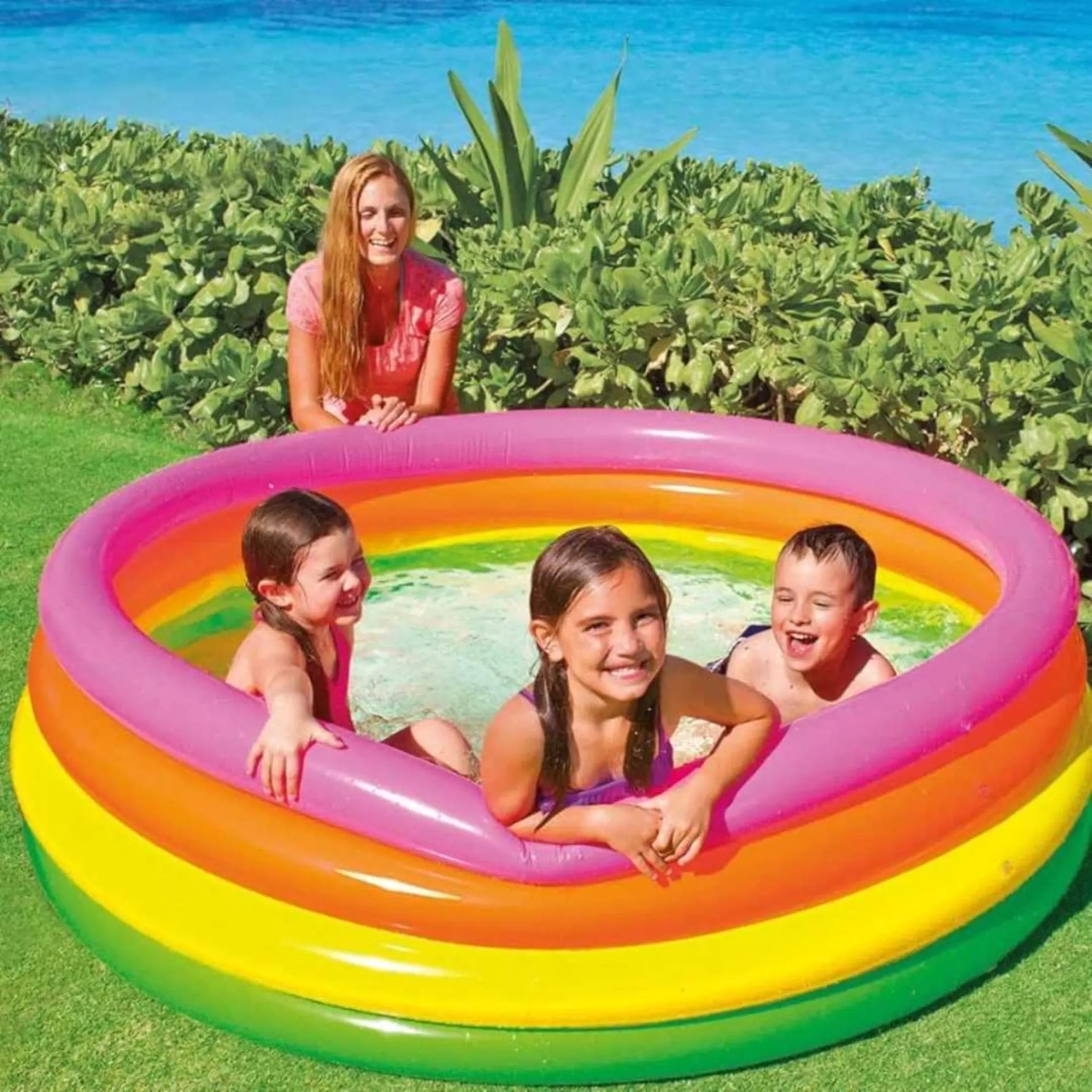 Children playing in a colorful inflatable pool with a woman standing behind them, surrounded by greenery.