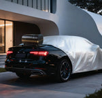 A black sedan parked outdoors in front of a modern building, with a white car cover draped over the vehicle and a black hardtop trunk lid mounted on top.
