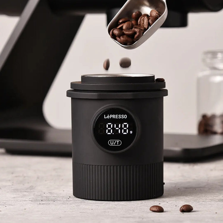 Black coffee grinder with digital display and coffee beans being added, set against a blurred kitchen background.