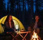 Two people sitting by a campfire at night in a forest, with a tent in the background.