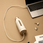White water bottle with charging cable connected to a laptop on a brown surface.