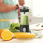 Portable blender with green smoothie surrounded by oranges, bananas, and spinach on a table.