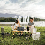 Two people sitting on a camping chair by a lake with outdoor equipment, including a portable stove and lantern.